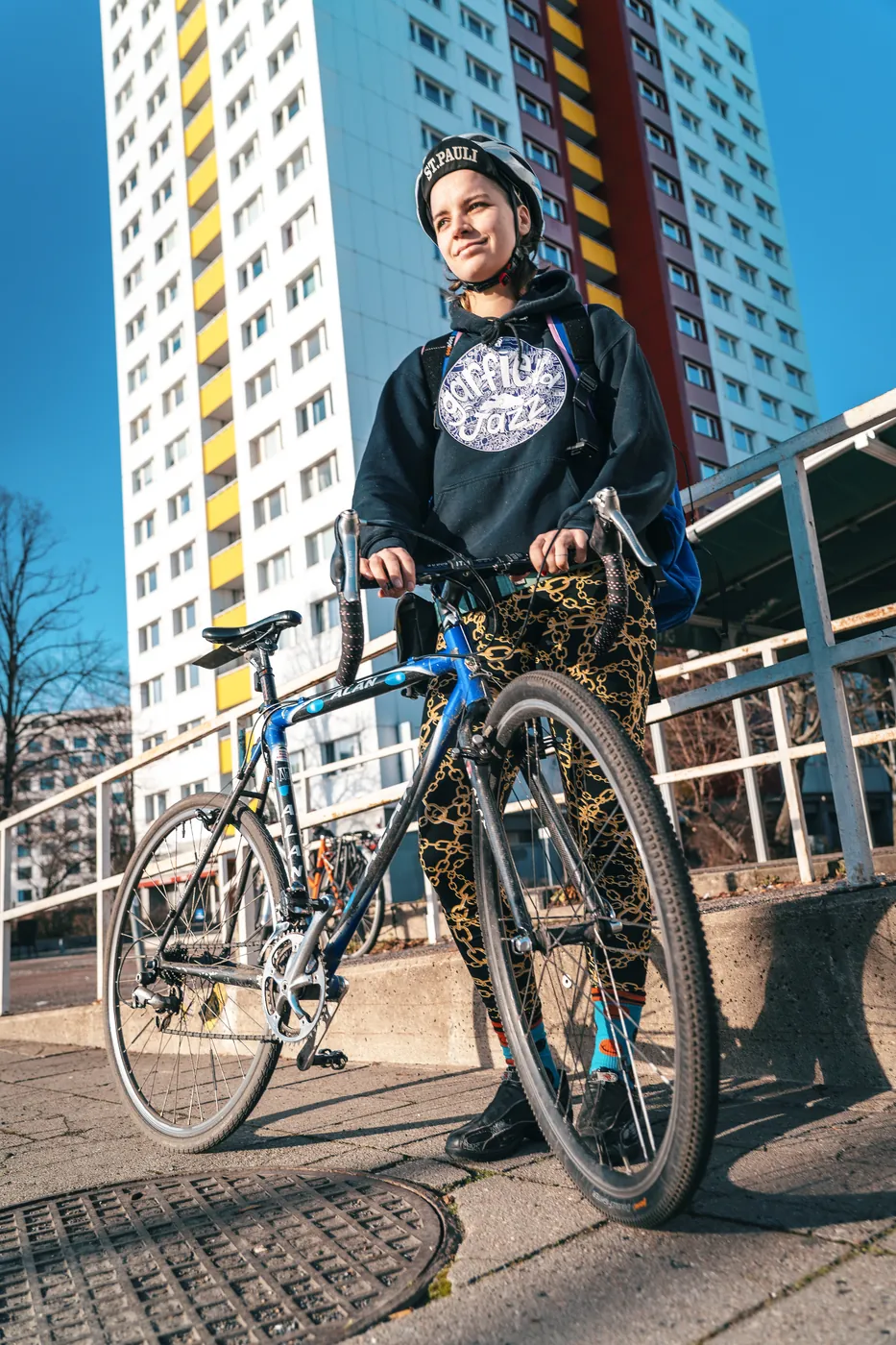 Khora courier holding a blue bicycle in front of a colorful Berlin apartment tower.